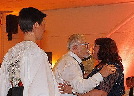 Ein Mann und eine Frau tanzen zusammen, lachen und lächeln. Eine weitere Person steht in der Nähe und beobachtet. Der Raum ist mit warmer orangefarbener Beleuchtung schwach beleuchtet.