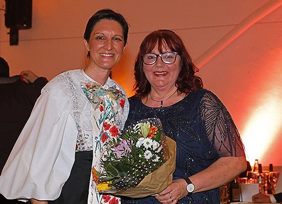Zwei lächelnde Frauen stehen zusammen und halten einen Blumenstrauß; eine trägt ein weißes Oberteil und die andere ein blaues Kleid mit einer Halskette. Hinter ihnen ist ein rotes Licht und Flaschen zu sehen.