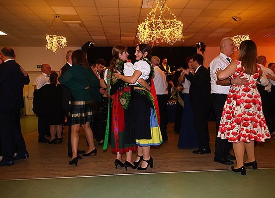 Eine Gruppe von Menschen tanzt in einem Saal mit Kronleuchtern. Einige tragen traditionelle Kleidung, während andere formelle Kleidung tragen. Eine Band spielt Musik im Hintergrund.