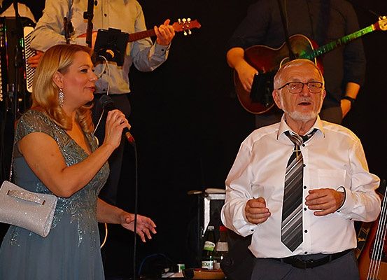Eine Frau in einem blauen Kleid singt in ein Mikrofon, während ein Mann in einem weißen Hemd neben ihr steht. Hinter ihnen spielen Musiker Gitarren und ein Akkordeon.