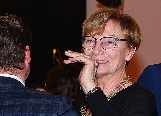 Eine ältere Frau mit Brille und Halskette lächelt und schaut jemanden vor sich an. Sie berührt ihr Kinn mit der Hand. Dahinter ist eine verschwommene Ansicht eines Mannes mit weißem Hemd und einer Person mit blauem Hemd.