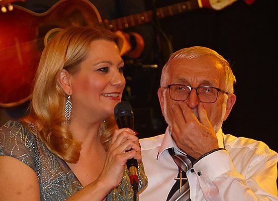 Eine Frau mit blonden Haaren und einem Mikrofon, lächelnd, steht neben einem älteren Mann mit Brille, der seinen Mund bedeckt. Hinter ihnen ist eine Gitarre zu sehen.