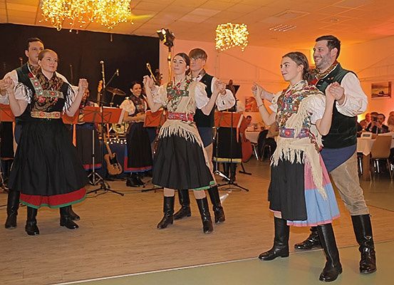 Fünf Personen tanzen auf einer Bühne, gekleidet in traditionellen Kostümen. Eine Band spielt hinter ihnen. Die Bühne ist mit Musikinstrumenten und Scheinwerfern geschmückt. Ein Publikum beobachtet im Hintergrund.