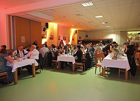 Ein Raum mit Tischen und Stühlen, viele Menschen sitzen und essen. Auf den Tischen befinden sich Flaschen, Gläser und Teller. Der Raum hat eine Projektionsleinwand an der Wand.