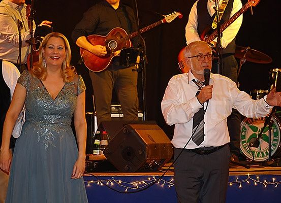 Ein Mann und eine Frau stehen mit einem Mikrofon und einem Lautsprecher auf der Bühne. Dahinter spielt eine Band auf der Bühne mit Gitarren, Trommeln und einer Becken.