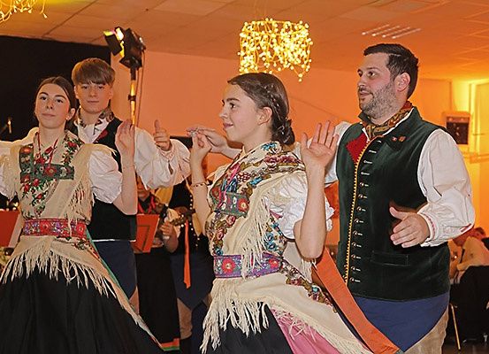 Vier Menschen in traditioneller Kleidung tanzen in einem Saal. Zwei Frauen und zwei Männer lächeln und halten Bänder. Dahinter spielt ein Musikorchester Instrumente.