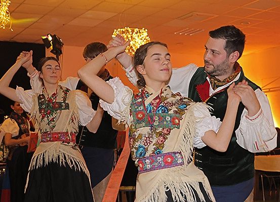 Eine Gruppe von Menschen in traditioneller Kleidung tanzt in einem Raum. Der Raum ist in orangem Licht gehalten.