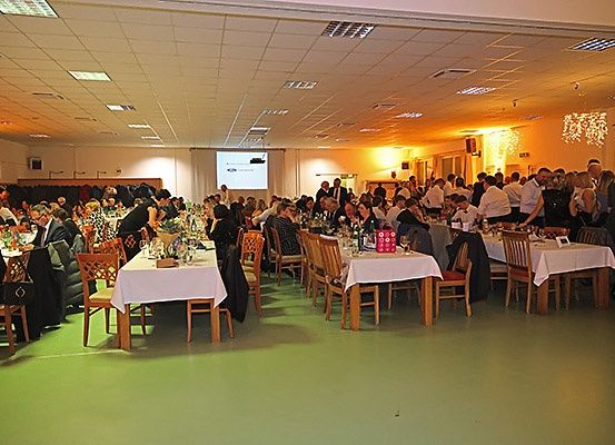 Ein Raum mit mehreren Tischen, die mit weißen Tischdecken bedeckt sind, besetzt von Menschen in formeller Kleidung. Eine Projektionsleinwand und Lautsprecher befinden sich an der Wand. Der Raum hat grünen Boden und eine weiße Decke.