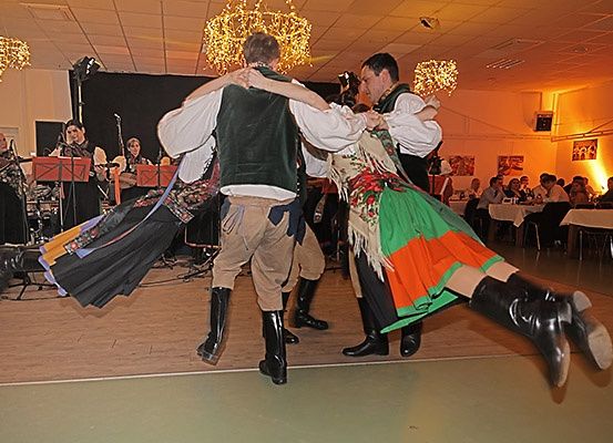 Drei Personen in traditionellen Kostümen tanzen auf einer Bühne, während Musiker hinter ihnen spielen und ein Publikum zuschaut.