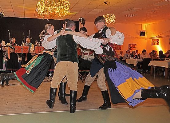 Vier Menschen in traditioneller Kleidung tanzen zusammen auf einer Bühne mit einer Band hinter ihnen, in einem Raum mit sitzenden Gästen und warmer Beleuchtung.