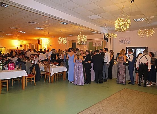 Eine große Gruppe von Menschen, die formell gekleidet sind, versammelt sich drinnen. Sie stehen und unterhalten sich, während Tische mit weißen Tischdecken und Stühlen sie umgeben. Dekorative Lichter hängen von der Decke.