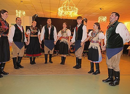 Eine Gruppe von Menschen in traditionellen Kostümen führt einen Tanz auf der Bühne auf. Sie halten sich an den Händen, lächeln und wenden sich dem Publikum zu.