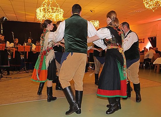 Eine Gruppe von Menschen in traditioneller Kleidung tanzt in einem Raum mit Holzboden. Eine Band spielt Musik im Hintergrund.