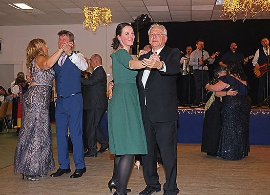 Mehrere Paare tanzen in einem Raum mit einer Bühne. Eine Band spielt hinter einem blauen Hintergrund. Der Raum hat Holzböden und ist mit Lichtern dekoriert.