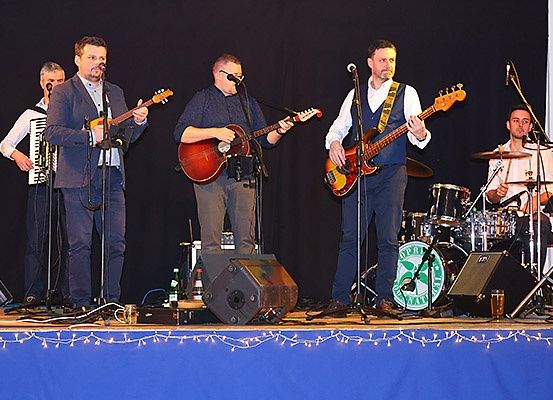 Eine Band tritt auf der Bühne auf, einer spielt Gitarre, ein anderer Bass und ein Dritter Schlagzeug. Ein viertes Mitglied spielt Akkordeon. Die Bühne ist mit Lichtern dekoriert.
