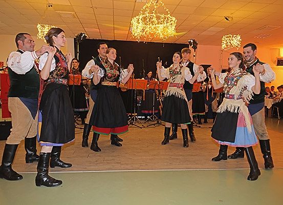 Eine Gruppe von Menschen in traditionellen Kostümen tanzt auf der Bühne vor einer Band, mit Kronleuchtern, die von der Decke hängen.