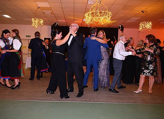Eine Gruppe von Menschen tanzt in einem Ballsaal. Einige sind in formeller Kleidung. Eine Kronleuchter hängt von der Decke.