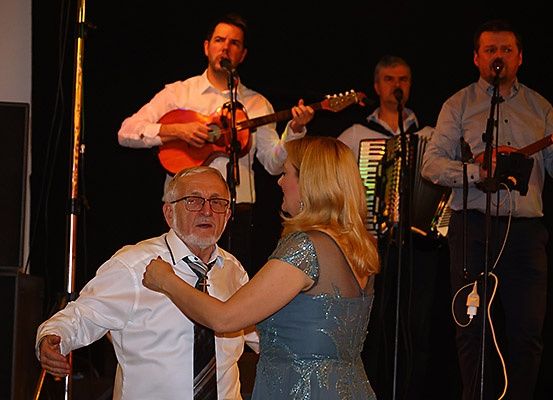 Ein älterer Mann und eine Frau tanzen auf einer Bühne, während eine Band hinter ihnen spielt. Die Bandmitglieder spielen Gitarre und Akkordeon. Die Frau trägt ein blaues Kleid und der Mann ein weißes Hemd. Sie lächeln und genießen die Aufführung.