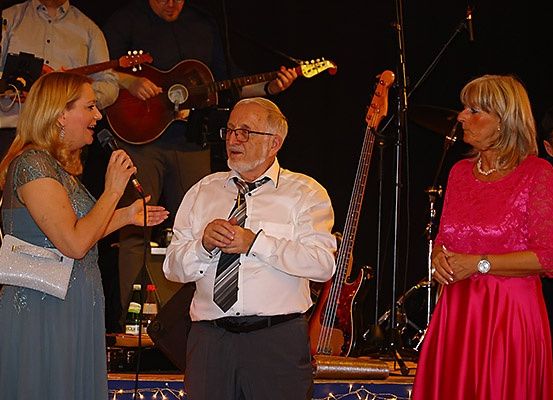 Drei Personen stehen auf der Bühne; eine Frau hält ein Mikrofon, eine andere Frau steht hinter einem Mann mit einer Gitarre im Hintergrund.