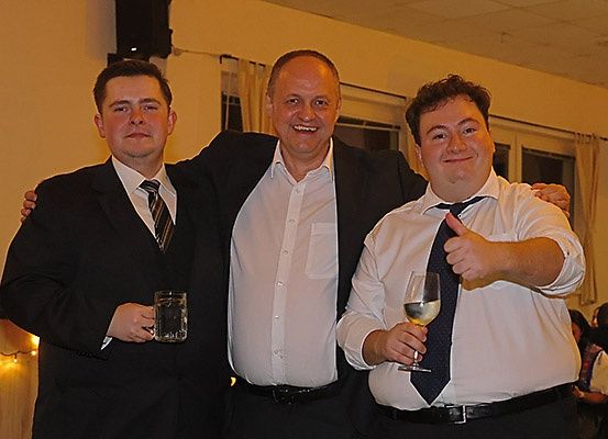 Drei Männer stehen in formeller Kleidung nebeneinander und lächeln für die Kamera. Der Mann auf der linken Seite hält ein Glas Bier, der Mann in der Mitte hat ein Glas Wein und der Mann auf der rechten Seite gibt einen Daumen hoch. Hinter ihnen sitzen und stehen weitere Personen, und links ist ein Lautsprecher zu sehen.