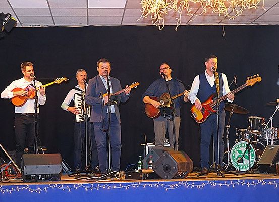 Fünf Männer spielen auf der Bühne mit verschiedenen Instrumenten, darunter Gitarren, ein Akkordeon und ein Schlagzeug. Sie singen und spielen alle zusammen.