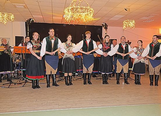 Eine Gruppe von Menschen in traditionellen Kostümen, die sich an den Händen halten, tritt auf einer Bühne auf, mit Musikern im Hintergrund.
