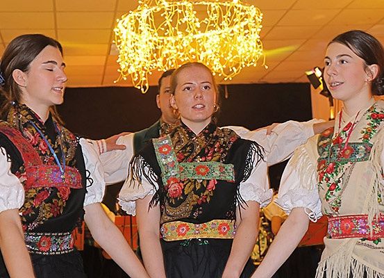 Drei Frauen in traditioneller Kleidung stehen lächelnd nebeneinander. Hinter ihnen hält ein Mann ihre Arme. Sie befinden sich in einem Raum mit einem Kronleuchter.