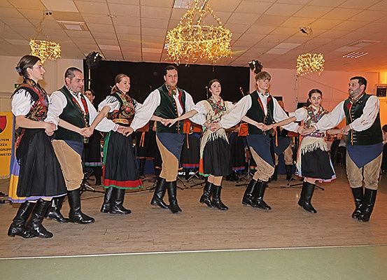 Eine Gruppe von Tänzern in traditionellen Kostümen tritt auf einer Bühne mit Holzboden auf. Sie halten sich an den Händen und scheinen mitten in einem Tanz zu sein.