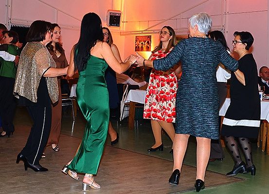 Eine Gruppe von Frauen tanzt in einem Partysaal. Sie tragen elegante Kleider und hohe Schuhe. Es gibt Tische mit weißen Tischdecken, und einige Flaschen stehen darauf.
