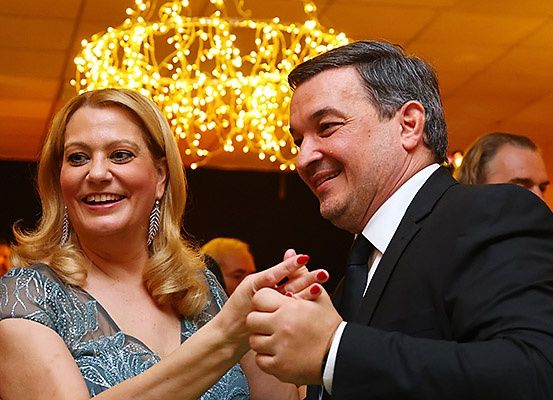 Ein Mann und eine Frau tanzen bei einer Veranstaltung, halten Händchen und lächeln. Die Frau hat blonde Haare und trägt ein blaues Kleid. Dahinter hängt ein Kronleuchter mit gelben Lichtern.