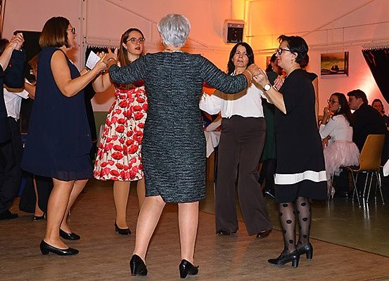 Eine Gruppe von Frauen tanzt zusammen in einem Raum mit gedämpfter Beleuchtung. Eine Frau hält ein Papier und sie tragen alle formelle Kleider.