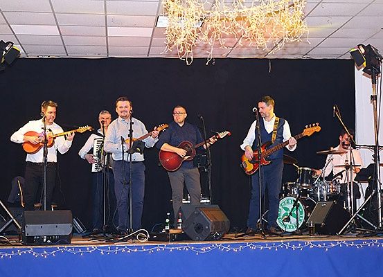 Eine Band spielt auf der Bühne mit Gitarren, Akkordeon und Schlagzeug. Sie stehen unter Scheinwerfern mit Lautsprechern und einer darüber hängenden Kristallleuchte.