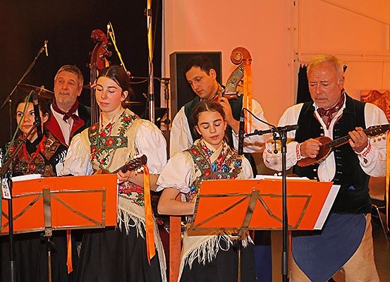 Eine Gruppe von Musikern in traditionellen Kostümen spielt auf der Bühne. Sie halten Gitarren und stehen vor Musikständern mit orangefarbenen Blättern. Dahinter spielt ein Mann Cello.