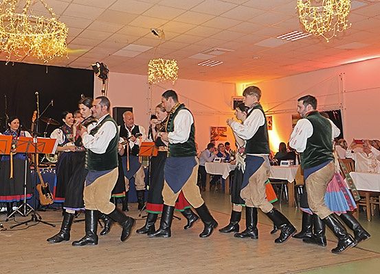 Eine Gruppe von Menschen in traditionellen Kostümen führt einen Tanz auf einer Bühne mit Musikinstrumenten und Kronleuchtern im Hintergrund auf.