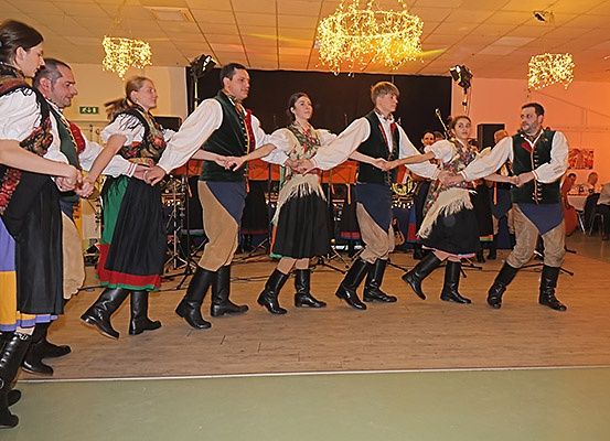 Eine Gruppe von Menschen in traditioneller Kleidung tanzt in einem Raum mit Holzboden und Kronleuchtern.