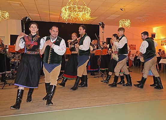 Mehrere Personen in traditionellen Kostümen tanzen auf einer Bühne, während im Hintergrund Musikinstrumente zu hören sind.