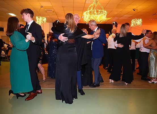 Eine Gruppe von Menschen tanzt in einem Raum mit Kronleuchtern. Einige tragen formelle Kleidung, während andere Freizeitkleidung tragen. Der Raum hat einen Holzboden und orangefarbene Beleuchtung.