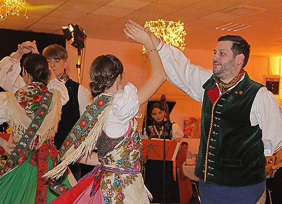 Ein Mann in traditioneller Kleidung führt zwei Frauen in Volkstrachten an, sie tanzen, während eine Frau in ein Mikrofon auf der Bühne singt.