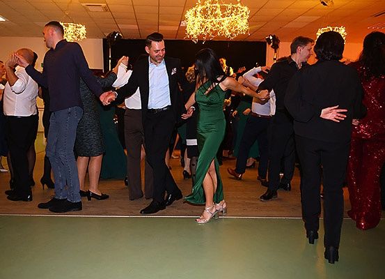 Eine Gruppe von Menschen tanzt zusammen in einem Saal. Einige halten sich an den Händen und lächeln. Eine Krone leuchtet von der Decke.