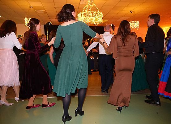 Eine Gruppe von Menschen tanzt in einem Ballsaal, gekleidet in elegante Abendgarderobe. Eine Frau in einem grünen Kleid führt den Tanz an.