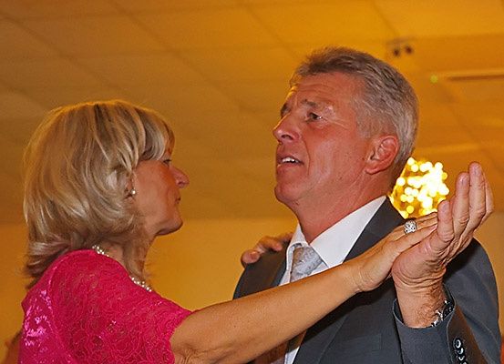 Ein älteres Paar tanzt auf einer Party. Die Frau trägt ein rosa Kleid und der Mann einen Anzug. Sie halten Händchen und schauen sich an. Eine Kronleuchter hängt von der Decke.