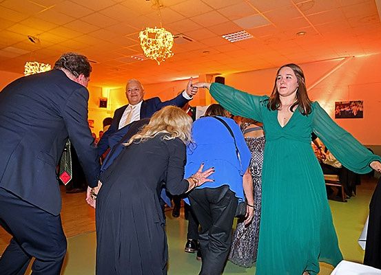 Mehrere Menschen tanzen in einem Raum mit schwacher Beleuchtung. Eine Frau in einem grünen Kleid zeigt auf etwas, während ein Mann im Anzug gestikuliert. Andere Menschen tanzen ebenfalls.