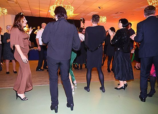 Menschen tanzen in einem Raum mit Holzboden und Kronleuchtern, in formeller Kleidung. Ein Mann und eine Frau stehen sich gegenüber und klatschen in die Hände.