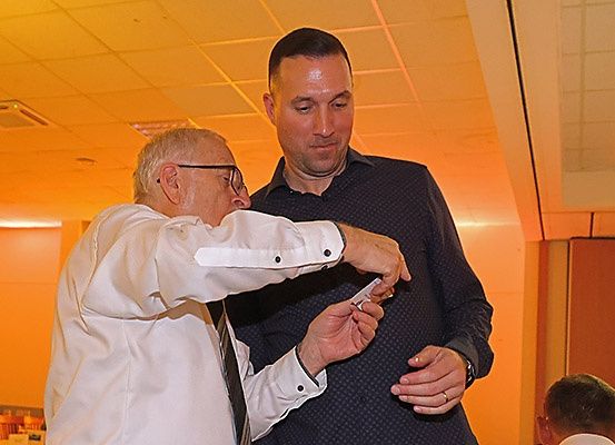 Zwei Männer stehen in einem Raum, einer zeigt dem anderen etwas, der schaut ihn an. Der Mann, der etwas zeigt, trägt eine Uhr und einen Ring.
