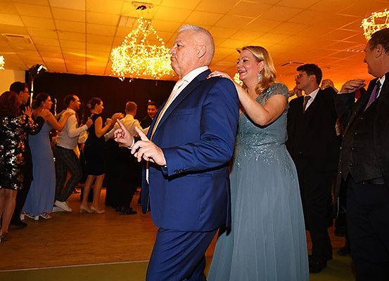 Eine Gruppe von Menschen tanzt in einem Raum mit Kronleuchtern. Ein älterer Mann und eine Frau in blauen Kleidern tanzen in der Mitte. Andere stehen herum und beobachten sie.