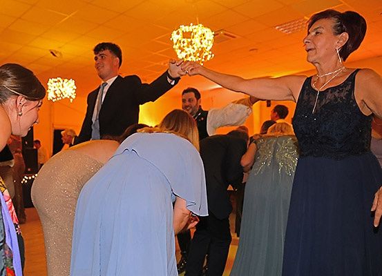 Eine Versammlung von Menschen in einem Saal, mit einer Frau in einem schwarzen Kleid, die einen Tanz leitet, einem Mann in Anzug und Krawatte, der lächelt, und anderen, die den Moment genießen.