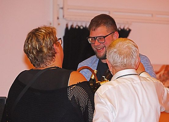 Drei Menschen stehen zusammen, zwei Männer und eine Frau. Die Frau hat kurze blonde Haare und trägt eine Brille. Die Männer lächeln und scheinen zu sprechen.