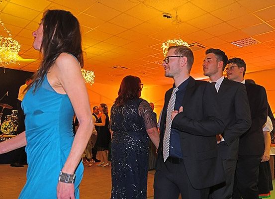 Eine Gruppe von Menschen tanzt in einem Saal mit gelbem Licht und einer Drum-Set im Hintergrund. Eine Frau in einem blauen Kleid und ein Mann in einem Anzug sehen sich an.