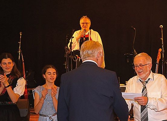 Eine Gruppe von Menschen steht auf der Bühne, zwei Frauen klatschen, ein Mann liest und ein weiterer steht mit einem Mikrofon hinter ihnen. Im Hintergrund sind Musikinstrumente zu sehen.