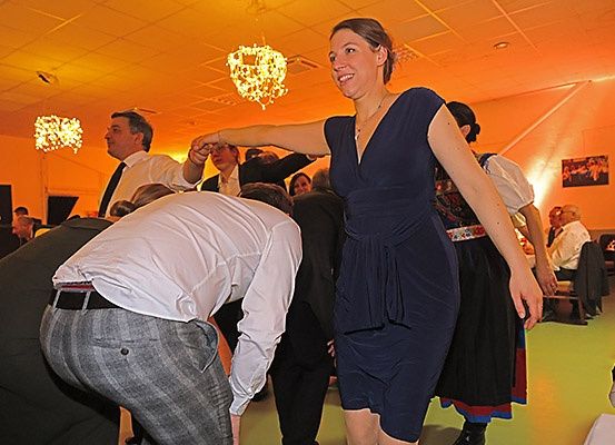 Eine Frau in einem blauen Kleid tanzt mit einem Mann in einem weißen Hemd auf einer Party. Hinter ihnen sitzen mehrere Personen an Tischen, und Kronleuchter hängen von der Decke.